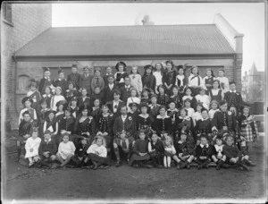 Large unidentified group of young girls, boys, two men and a woman outside a building, probably Christchurch district, shows most of them wearing Scottish national costume