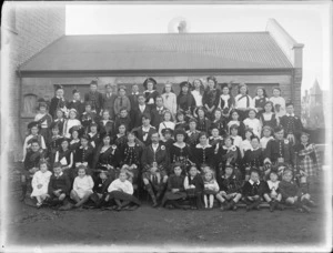 Large unidentified group of young girls, boys, two men and a woman outside a building, probably Christchurch district, shows most of them in Scottish national costume