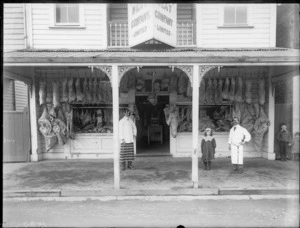 Exterior view of a butcher shop, showing two unidentified butchers and small children standing outside the store with carcasses hanging from the ceiling, probably Christchurch district