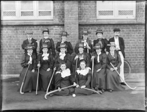 Group of unidentified female hockey players wearing hats [with letters LHC?] outside a brick building, including two men [coaches?], probably Christchurch district
