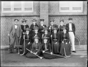 Group of unidentified female hockey players wearing hats [with letters LHC?] outside a brick building, including two men [coaches?], probably Christchurch district