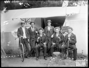 Group of six unidentified young men wearing straw boaters outside a tent hung with union Jack flags, sign behind them reads 'Myrtle Camp' and a sign in front reading 'Please Keep Off The Grass', [New Brighton, Christchurch district?]