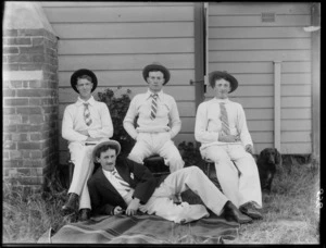 Four unidentified young men wearing hats, three sitting in chairs, one lying in front, on the grass outside a wooden building with brick fireplace, with a dog alongside, probably Christchurch region