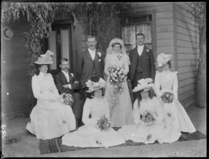 Wedding party portrait by front door of wooden house, unidentified bride with veil, groom and two best men with lapel flowers, four bridesmaids including twins, women in hats holding flowers, probably Christchurch region
