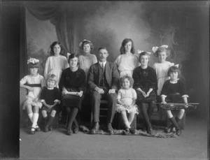 Group portrait taken in a studio, showing a man with 10 girls [daughters?], one of whom holds a violin, and another a rolled-up document, Christchurch