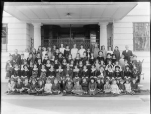 Large group of girls dressed in traditional Scottish [dancing?] costume, including some decorated with medals, flanked by bagpipers and unidentified adults and children, outside entrance to large stone building, possibly Christchurch district