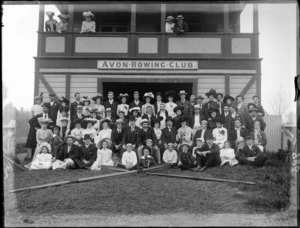 Avon Rowing Club building, with unidentified men and women club members on balcony, and with children on grass slope in front, wearing an assortment of hat and caps, with rowing oars and rudder in front, probably Christchurch region