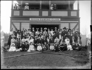 Avon Rowing Club building, with unidentified men and women club members on balcony, and with children on grass slope in front, wearing an assortment of hat and caps, with rowing oars and rudder in front, probably Christchurch region