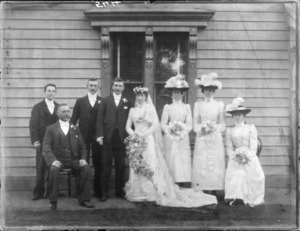 Wedding group, including bridesmaids wearing hats adorned with ostrich feathers, all members unidentified, outside a wooden house, possibly Christchurch district