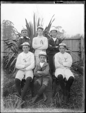 Portrait of six unidentified young boy coxswains in uniforms and white caps with 'cox' and Canterbury Rowing Club emblems, on long grass in front of a flax bush and wooden fence, probably Christchurch region