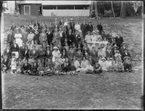 Large group portrait of unidentified men, women and children on a social outing, grass slope with wooden horse stables with people behind, probably Christchurch region