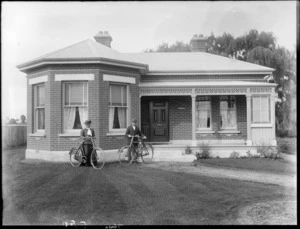 Exterior of a brick house, showing an unidentified man and woman with bicycles standing on front lawn, possibly Christchurch district