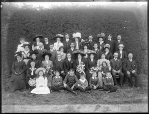 Large wedding group portrait on long grass with hedge beyond, unidentified bride and groom with extended family, women with hats, pageboy in sailor uniform, probably Christchurch region
