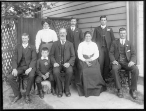 Family portrait, unidentified older couple with family members, four men, a woman, and a boy, with wooden lattice fence and house behind, probably Christchurch region