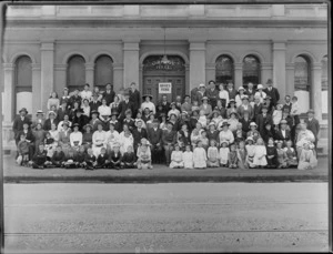 Large group portrait of unidentified men and women in hats with children, with Bruce's Picnic sign, outside the Orange Hall building of the Loyal Orange Institution charitable organisation, Christchurch District Number 5, probably Christchurch region