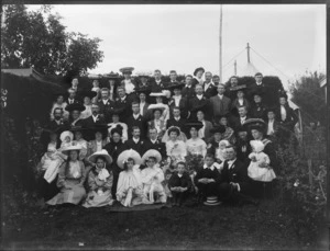 Wedding group, showing unidentified men, women and children, in an outdoor location, with tents visible above hedge behind, possibly Christchurch district