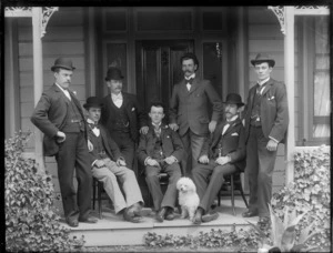 Group of unidentified men wearing business attire and bowler hats, standing with a small dog on a verandah at the front entrance to a wooden house, possibly Christchurch district
