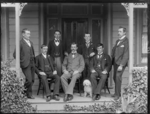 Group of unidentified men, without hats, wearing business attire, standing with a small dog on a verandah, at the front entrance to a wooden house, possibly Christchurch district