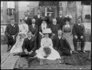 Wedding group portrait on grass in front of wooden house, unidentified bride and groom with parents and older family members, women in hats, probably Christchurch region