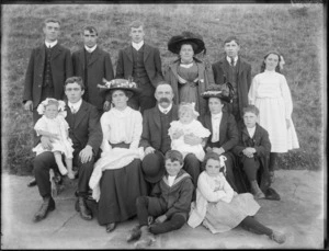 Family portrait on footpath in front of grass slope, unidentified older parents with adult family members and grandchildren, women in hats, probably Christchurch region