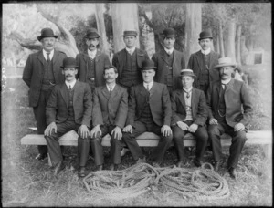 Group portrait of unidentified men in suits with bowler hats, on grass with gum trees behind, with coils of hawser laid rope in front [tug-of-war team?], probably Christchurch region