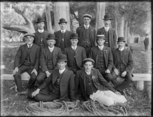 Group portrait of unidentified men in suits with bowler hats, on grass with gum trees behind, with coils of hawser laid rope in front [tug-of-war team?], probably Christchurch region