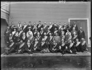 Large group portrait of mostly older unidentified men and a few women wearing embroidered sashes [Hibernian Australasian Catholics Benefit Society?], man with chain of office near front, in front of a wooden building, probably Christchurch region