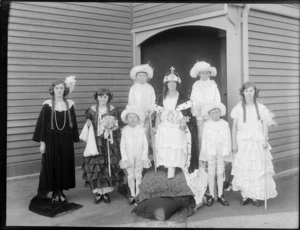 Group in fancy dress, showing a woman dressed as a queen, sitting on a makeshift throne, surrounded by four boy 'pages', and three girls [ladies-in-waiting?], outside a wooden [hall?] building, possibly Christchurch district