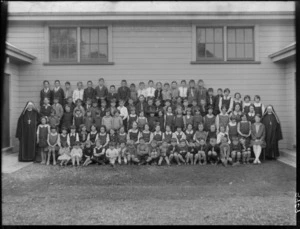 School class portrait in front of large wooden building, large number of unidentified children of various ages and two Catholic Nuns wearing crucifixes, probably Christchurch region