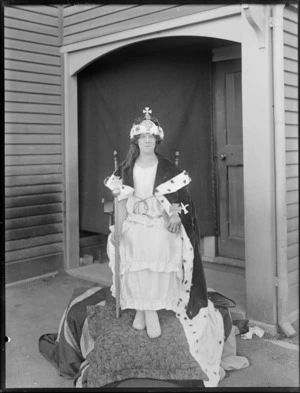 A woman, unidentified, dressed in fancy dress as a queen, sitting on a makeshift throne, outside an entrance to a wooden [hall?] building, possibly Christchurch district