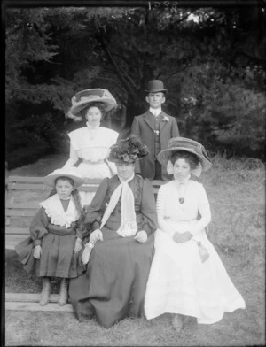 Family group, including two women wearing very large straw hats, a girl, and a man in a bowler hat, all unidentified, on a park bench, possibly Christchurch district