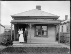 Exterior view of a wooden cottage, showing an unidentified man and woman with baby, on verandah, with the house number '46' visible behind, possibly Christchurch district