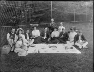Group picnicking, showing unidentified men, women and children sitting around food and crockery which is laid out on a cloth, in a grassed area enclosed by wire netting [tennis club?], possibly Christchurch district