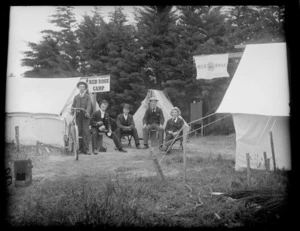 Five unidentified young men with hats, one with a bicycle, having a cup of tea, sitting between tents with 'Red Rose Camp' signs, trees beyond, [Sumner?], Christchurch