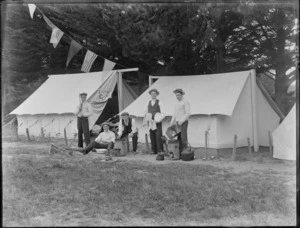 Five portrait young men with hats, two cooking over wood burners, in front of two tents with 'Tivoli Camp' sign and multiple flags, trees beyond, [Sumner?], Christchurch