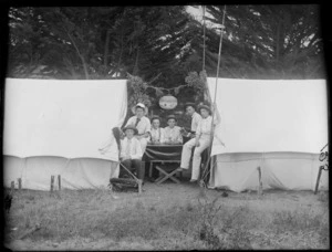 Six unidentified young men in hats sitting at a table between two tents with Chinese lantern above, one playing an accordion, trees beyond, [Sumner?], Christchurch