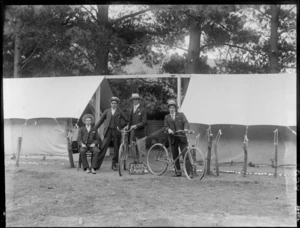 Four unidentified members of Flora Camp, one sitting and three standing between tents, with two holding bicycles, a sign for Flora Camp, probably Christchurch district