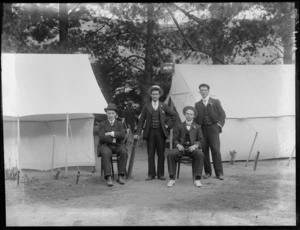 Four unidentified members of the Jubilee Camp, two sitting and two standing between tents, one showing a sign for Jubilee Camp, probably Christchurch district