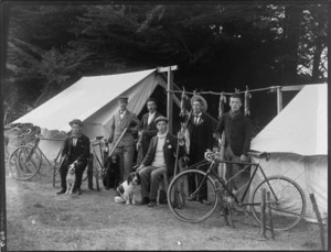 Rabbit-shooting party, showing six men with bicycles, guns, and dogs, standing beside two tents, which have rabbit carcasses strung between them, possibly Christchurch district