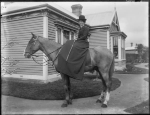 An unidentified woman dressed in riding habit sitting side saddle on a horse in front of a house, probably Christchurch district, showing a child in the window and a bicycle leaning against a tree in the background