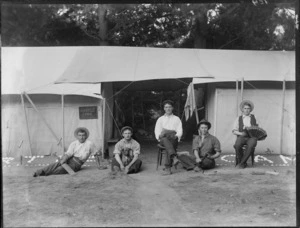 Five unidentified young men with hats and accordion, cricket bat, dog and gun, in front of two tents with awning and 'Myrtle Camp' sign, trees beyond, [Sumner?], Christchurch