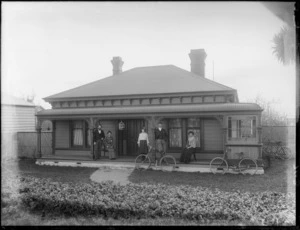 Members of an unidentified family on the front porch of the house, showing two women sitting on chairs, two men and a young girl standing and two bicycles on the front lawn, probably Christchurch district