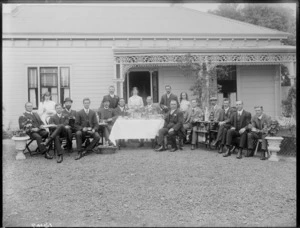 Outdoor tea party, showing a group of unidentified men and women, sitting on chairs around a table, which has been laid with food and flowers, with a wooden house behind, possibly Christchurch district