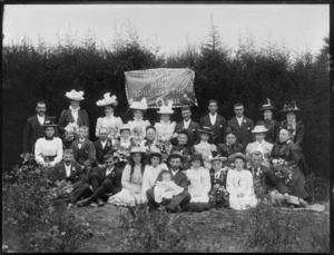 Bridal party, showing unidentified men, women and children with a banner behind them, reading 'Health to the bride, long live the happy pair', in a garden, possibly Christchurch district