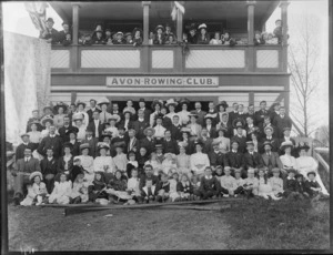Avon Rowing Club building, unidentified club members and children on balcony with British flag, and on grass slope in front with oars, wearing an assortment of hat and caps, Christchurch