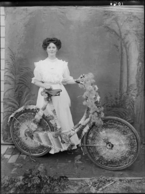 A full length portrait of an unidentified woman standing behind a decorated bicycle with crepe paper ribbons and a fluffy floral ribbon on the handle bars, taken in the studio, probably Christchurch district