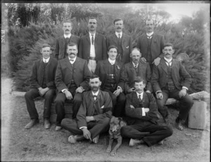Group of unidentified men in business attire, each with a small rosette pinned to lapel, some wearing a black armband, in an outdoor location, possibly Christchurch district