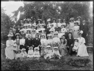 Large wedding group portrait, unidentified bride and groom with extended family, children and babies, women with hats, in the back garden with trees beyond, probably Christchurch region