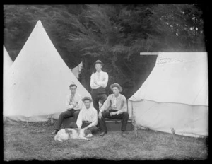 Four unidentified young men, with a dog, sitting next to tents, one of which has a sign reading 'Tawera Camp', possibly Christchurch district