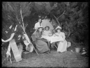 Group dining outdoors, showing two young women and two young men, all unidentified, sitting at a table, drinking tea, in a clearing enclosed by pine trees, with a tent visible in background, possibly Christchurch district